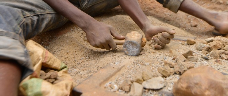A person sits on the ground with a mallet in his hand and rocks in front of him. Credit: Pact 